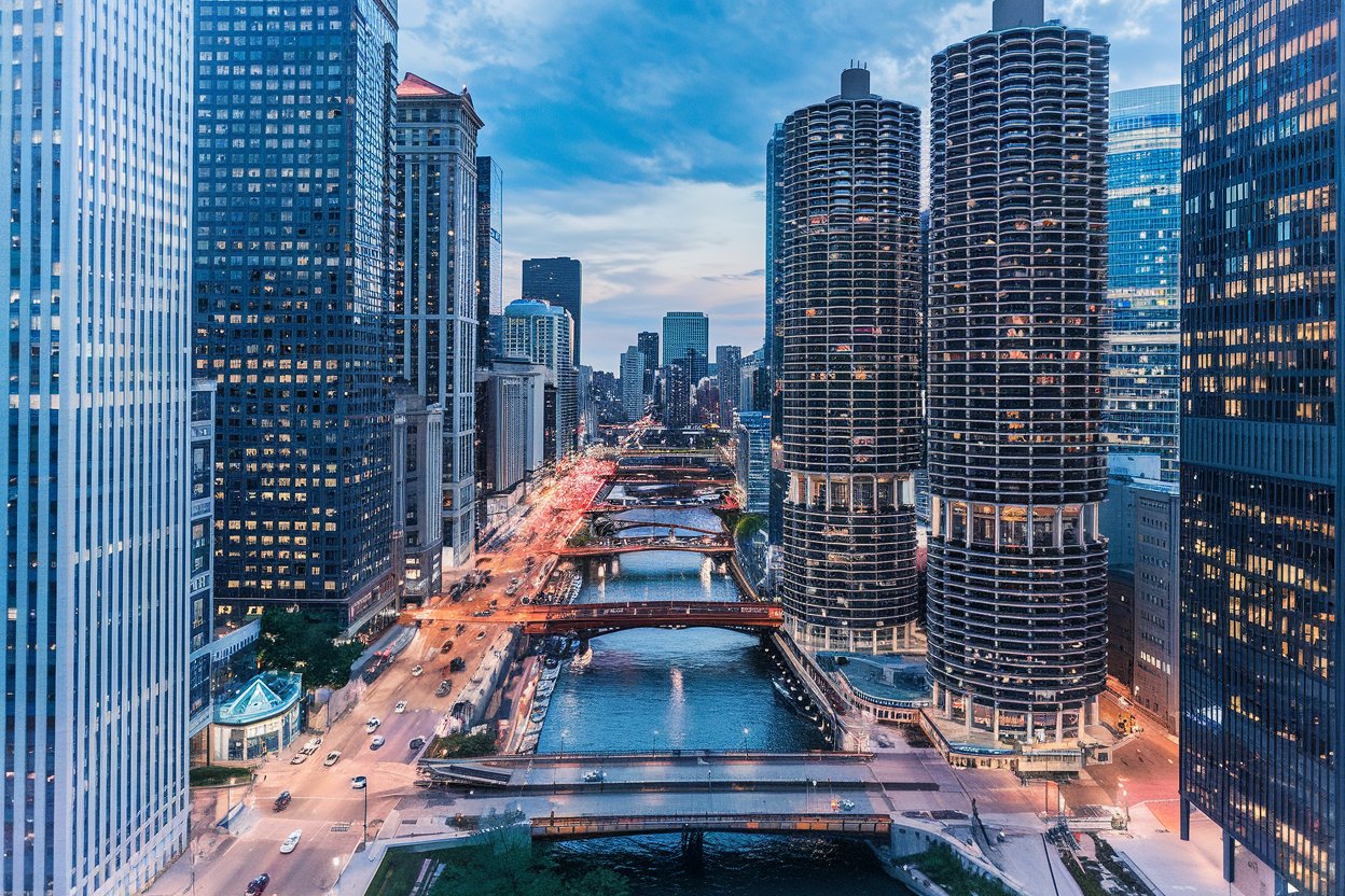 A lively scene along the Chicago Riverwalk, featuring waterfront cafes, lush greenery, and iconic skyscrapers reflected in the river.