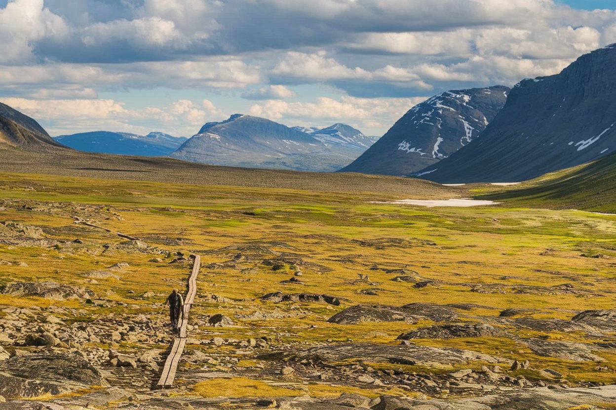 A hiker walking along the Kungsleden Trail in Swedish Lapland, surrounded by rugged mountains, pristine lakes, and vast wilderness.