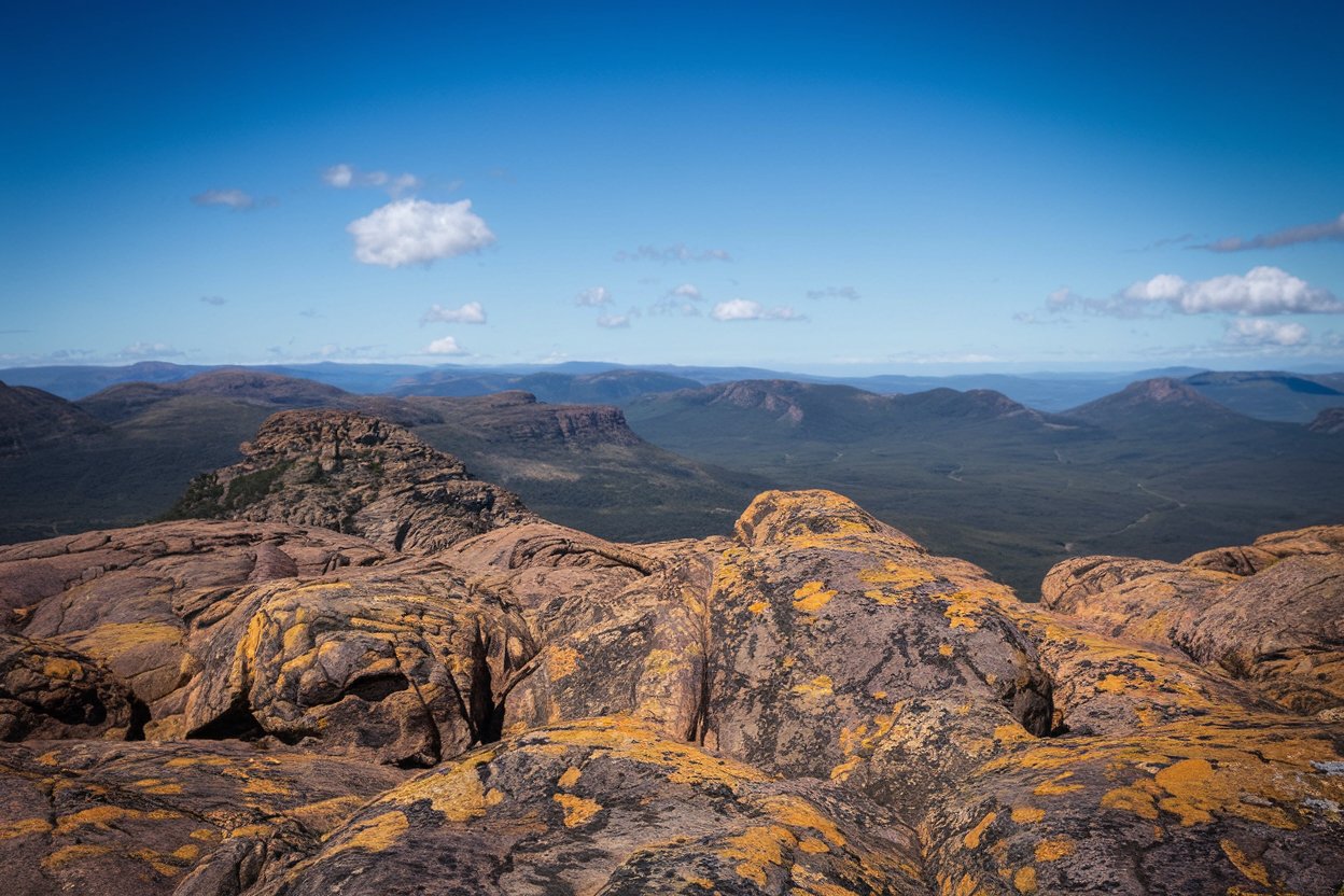 breathtaking landscape along the overland track in australia, with rugged mountains, expansive green valleys, orange-brown rocky formations, and a clear sky with scattered clouds