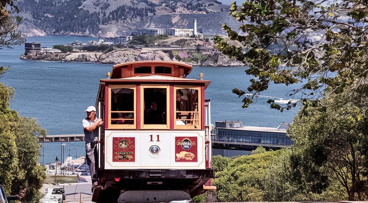 A historic San Francisco cable car climbing a steep street with the city skyline and bay in the background.